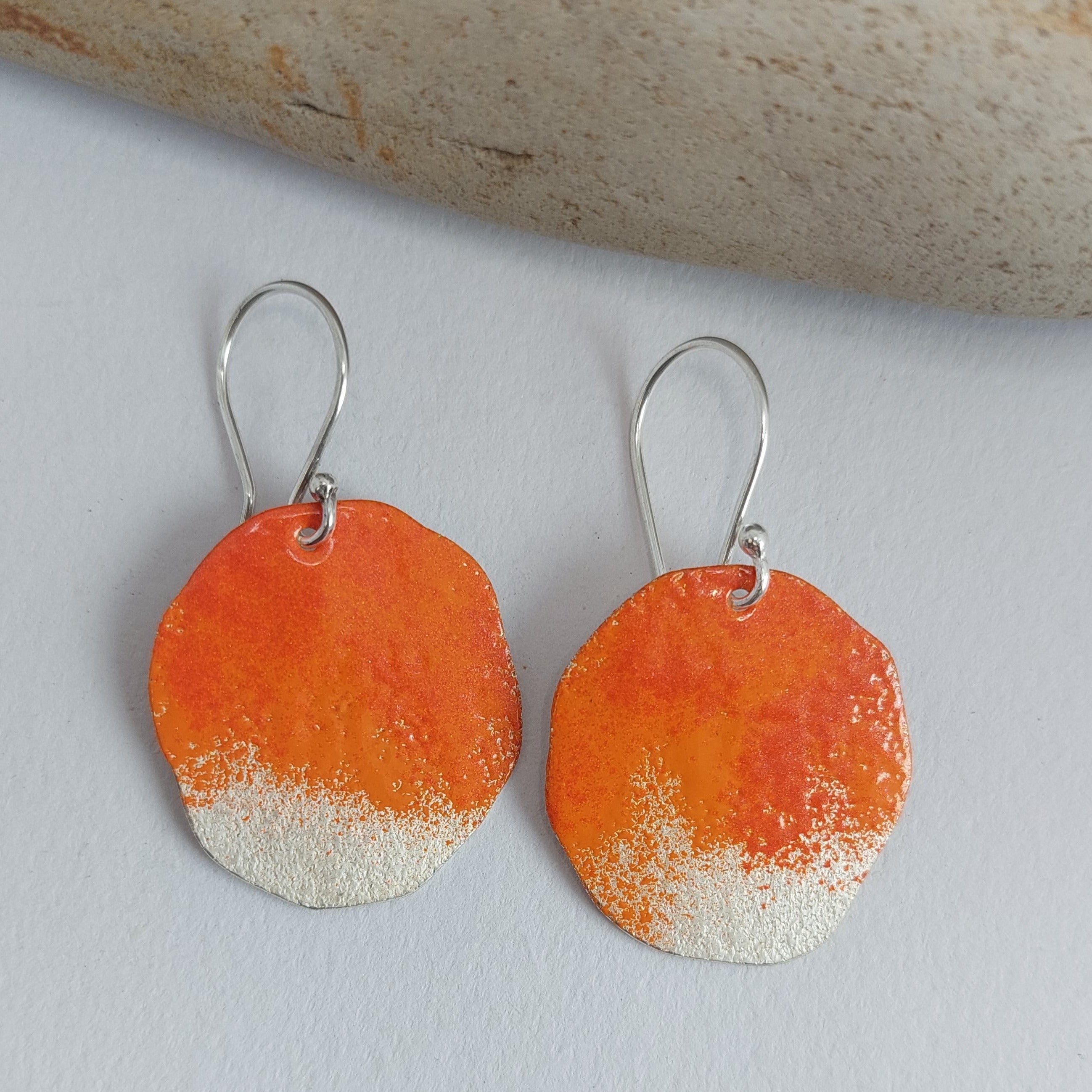 Organic shape sterling silver statement earrings with bright oramge powder coat paint on it on white background and a rock.