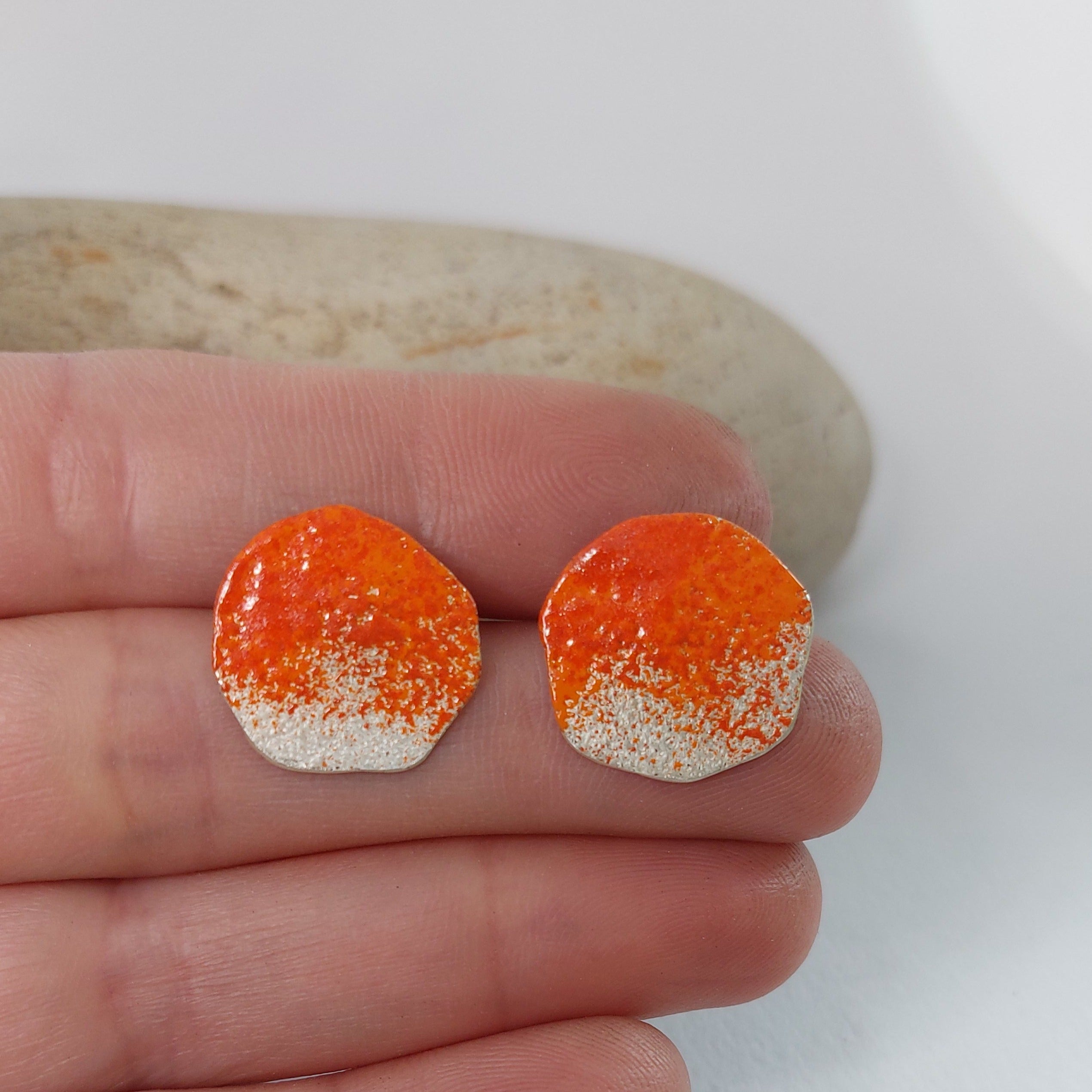 Sterling silver organic shape stud earrings with orange powdercoat held on a hand with a rock in the background.